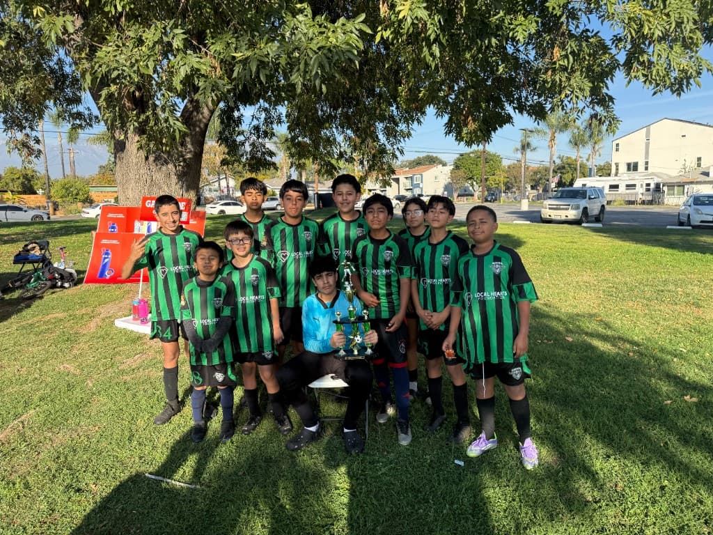Team victory portrait with the trophy, Local Hearts Foundation jerseys, and coaches on the pitch